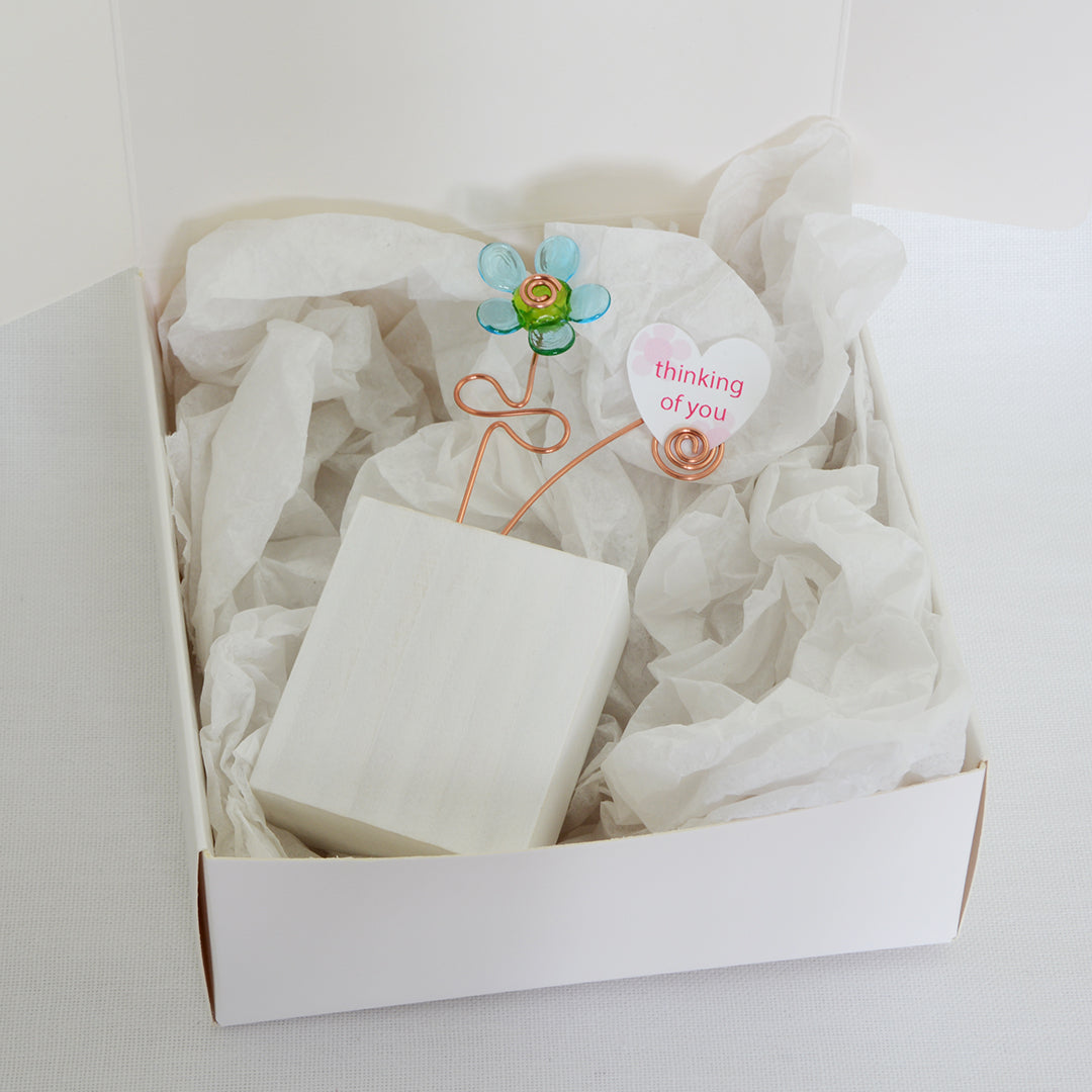 Turquoise and green glass flower on a white wooden block with a paper heart with the message 'Thinking of You' placed on tissue paper in a white gift box